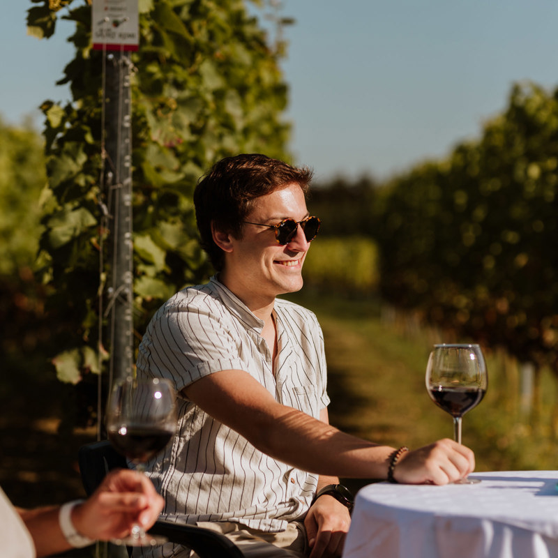Een lachende man zit aan een tafeltje tussen de wijngaarden van Raar Saint Remi met een rode wijn en een andere man