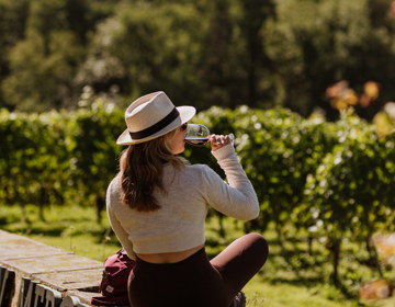 Een jonge vrouw drinkt uit een glas rode wijn terwijl ze op een muurtje zit en uitkijkt over de Zuid-Limburgse wijngaarden, heuvels en de kerk van Noorbeek