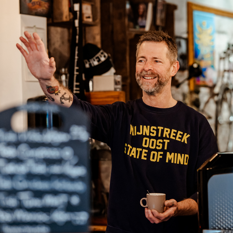 Patrick zwaait met koffie in hand in Pat's Tosti Bar
