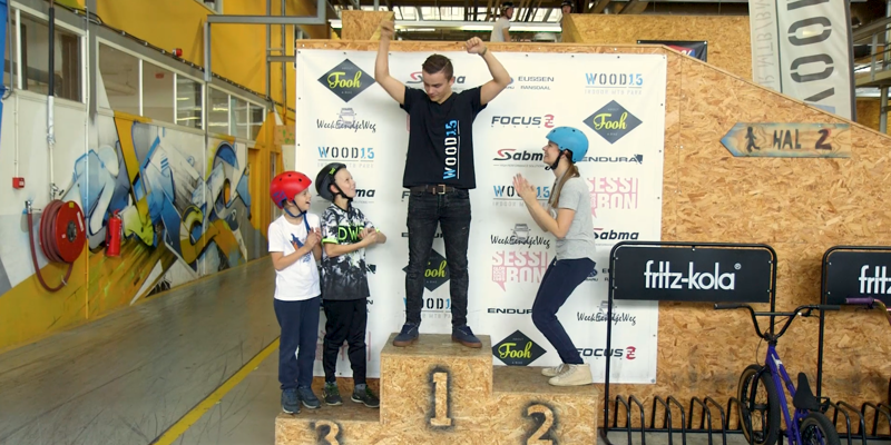 Jongen met handen in de lucht op 1e plek van podium. Kinderen en vrouw op plek 2 en 3 applaudiseren.