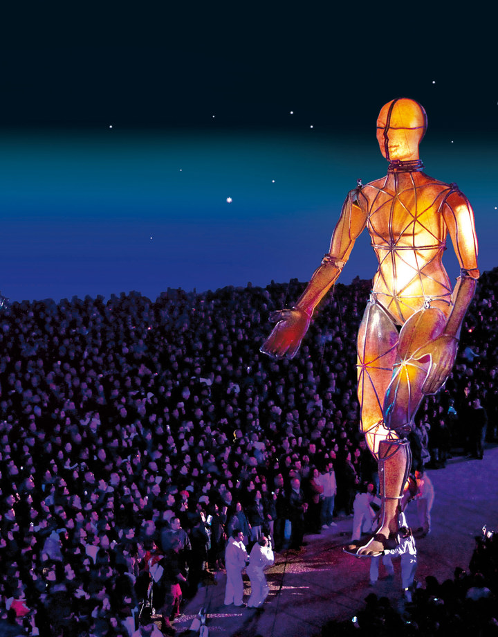 Enorm wandelend sculptuur van buizen en oranje licht loopt door menigte in het donker