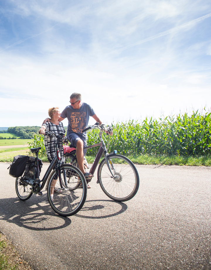 Een koppel op de fiets kijkt elkaar boven op de berg verliefd aan met achter zich een panorama over de heuvels