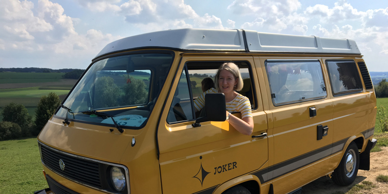 Vrouw zit achter het stuur van een geel camperbusje. 