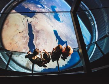 Een familie met twee kinderen staat op een glazen reling en kijkt uit over een grote interactieve wereldbol in het Discovery Museum