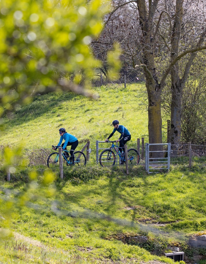 Twee wielrenners fietsen over een smal fietspaadje tussen de groene weilanden