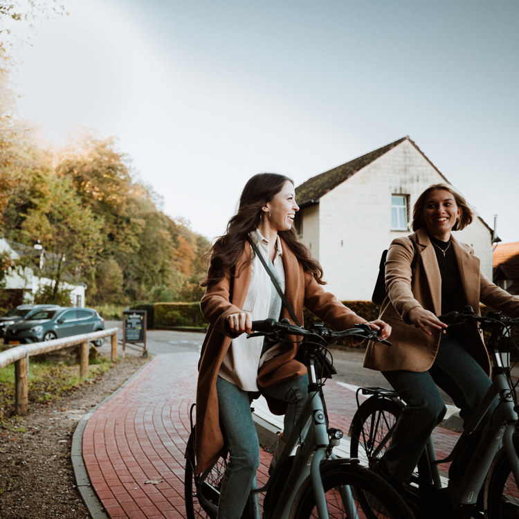 Twee dames Fietsen op een fietspad Langs het uithangbord van Brasserie De Groeve, langs een bosrand 