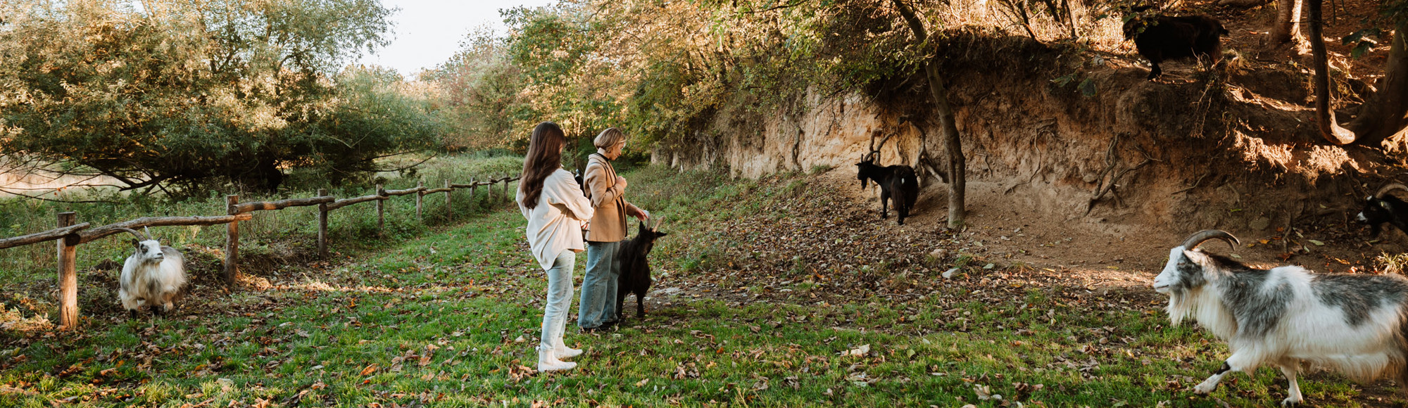 Twee vriendinnen staan in een wei met geitjes, met daarachter een heuvel van mergel en bomen