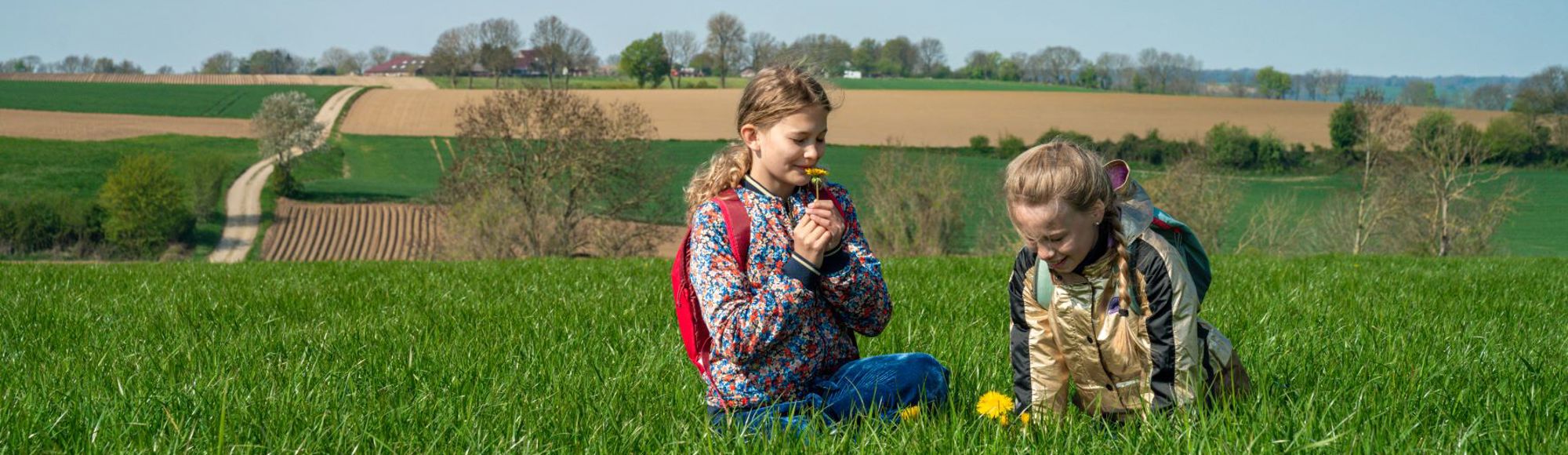 Twee blonde meisjes zitten in het gras en ruiken aan een bloem, met op de achtergrond het limburgse heuvelland. 