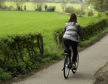 Een vrouw in een zilveren jas fiets over een fietspad met uitzicht over de Limburgse heuvels