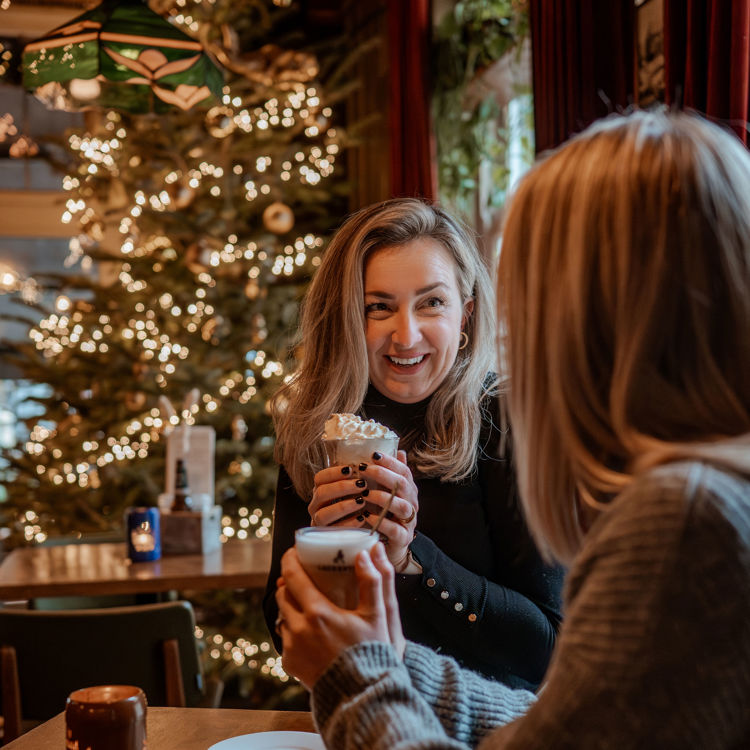 Twee vriendinnen zitten met warme chocomelk in een knus cafe tijdens de kerstperiode.