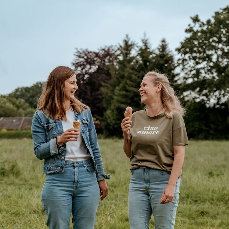 Twee dames lachen met een ijsje in de hand, in een natuurrijke omgeving.
