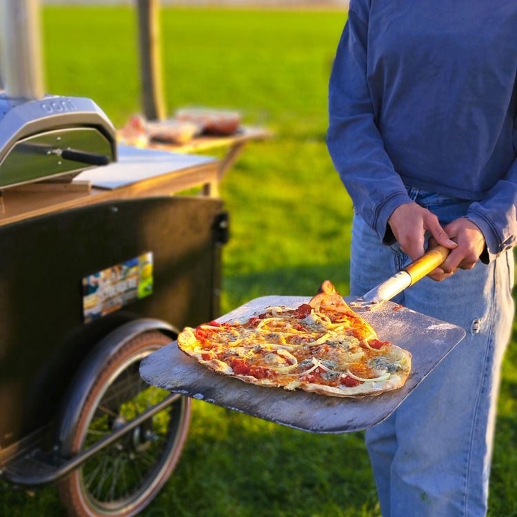 Meisje houd een verse pizza vast naast een pizzabakfiets. 
