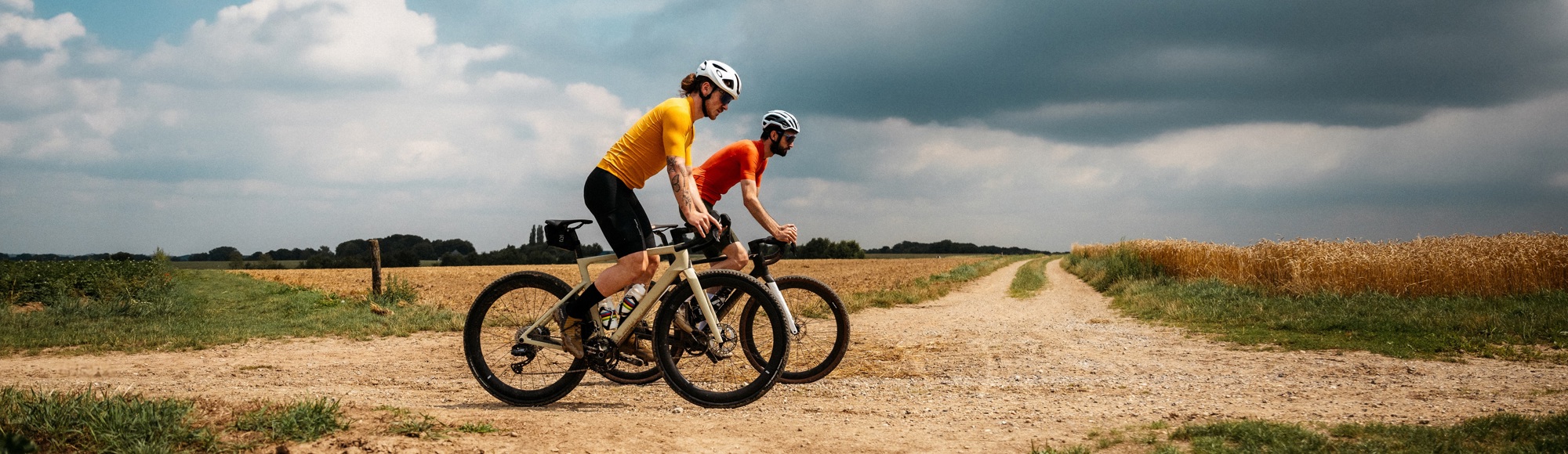 2 mannen met oranje fietspakjes op gravelbikes op onverharde weg tussen de velden
