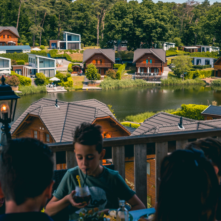 Gezin zit op balkon van vakantiehuisje aan het water, met nog meer vakantiehuisjes.