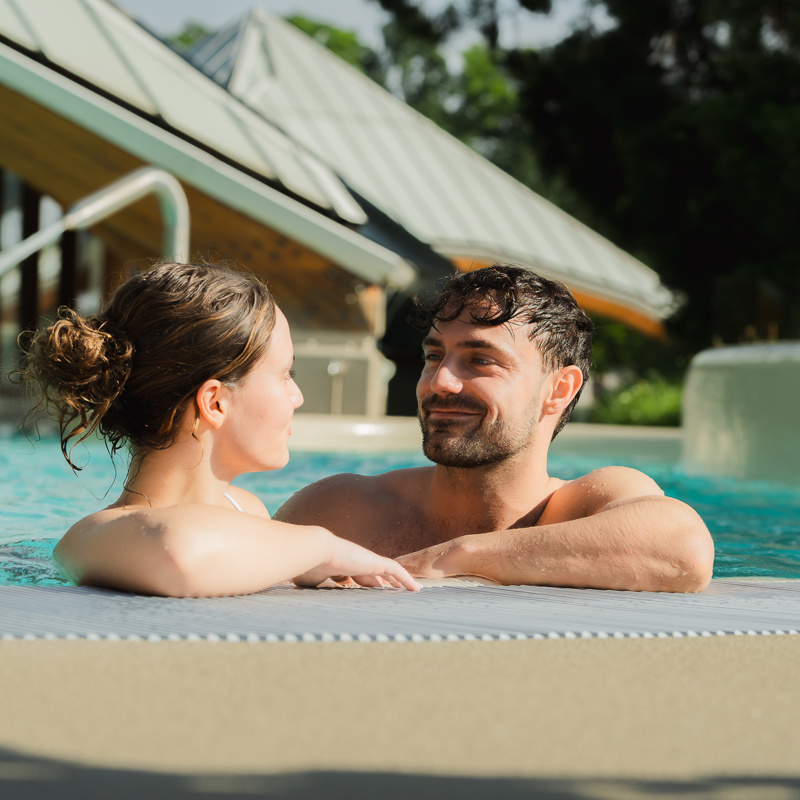 Jong koppel hangt over de rand van het thermale buitenbad van Thermae 2000.