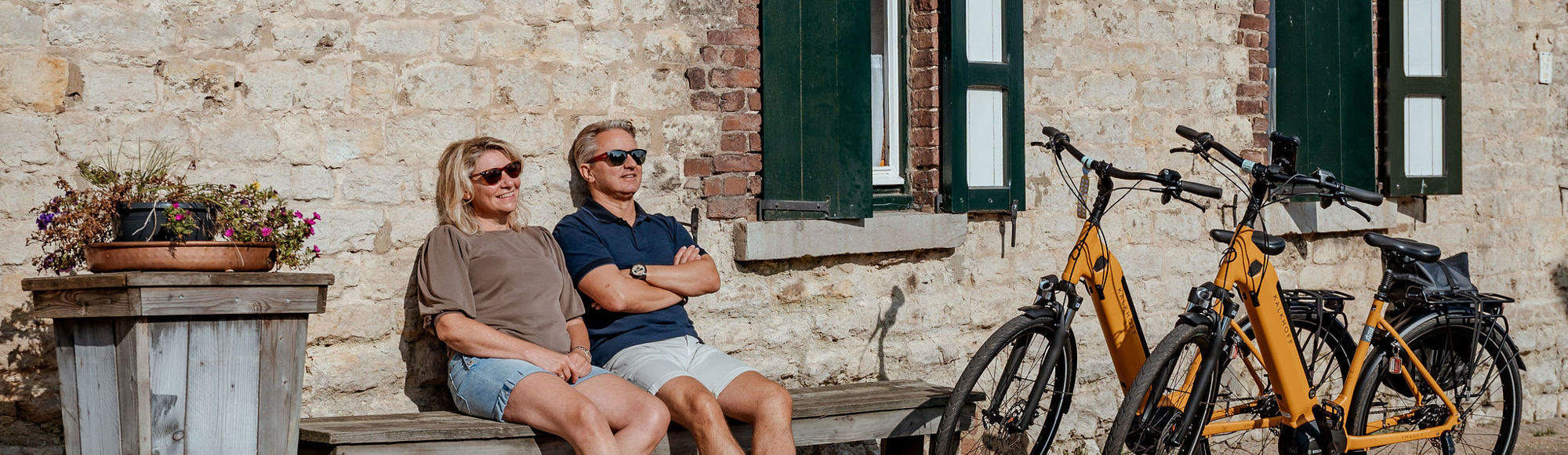 Koppel geniet tijdens fietstocht op een bankje van de zon en leunt tegen een mergelmuur.