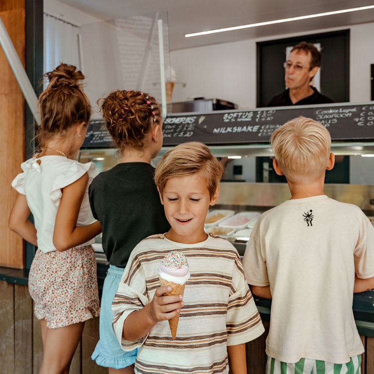 Jongetje kijkt blij naar zijn schepijsje met kinderen en een ijskraam op de achtergrond. 