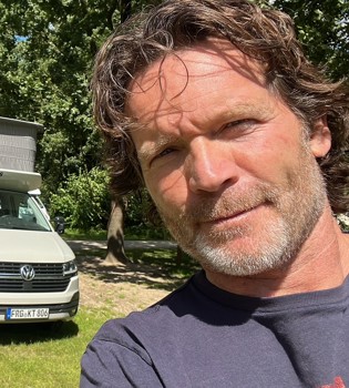 Selfie van Sander Janson op een camping met zijn kamper op de achtergrond. 